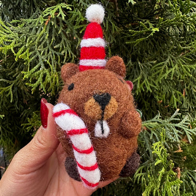 Small brown beaver felted ornament with a candy cane held in front of green foliage