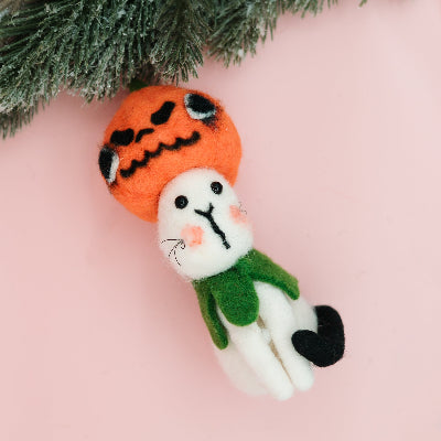 A small felt Halloween ornament shaped like a white cat wearing a green scarf and an orange pumpkin hat, photographed on a soft pink background with evergreen branches at the top.