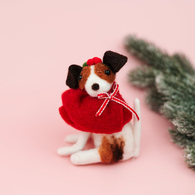 A small felt Christmas dog figurine wearing a red sweater and red-and-white striped bow, posed on a soft pink background with blurred greenery at the right edge.
