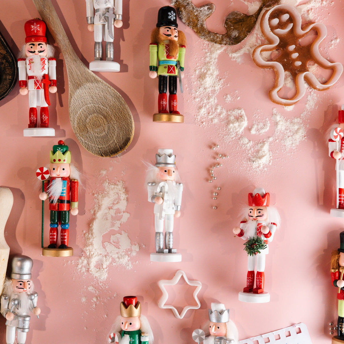 A styled flat-lay of assorted Christmas nutcracker figurines arranged on a pink background with baking props including a wooden spoon, rolling pin, flour, cookie cutters, and a gingerbread cookie.