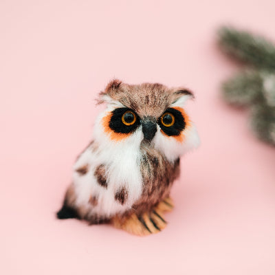 A small owl figurine with large round eyes, brown and white feathers, and orange accents around the eyes, sitting on a solid pink background with a blurred green pine branch in the upper right.