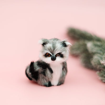 A small plush toy raccoon with gray, white, and black fur sitting on a solid pink background, with a blurred green pine branch on the right.